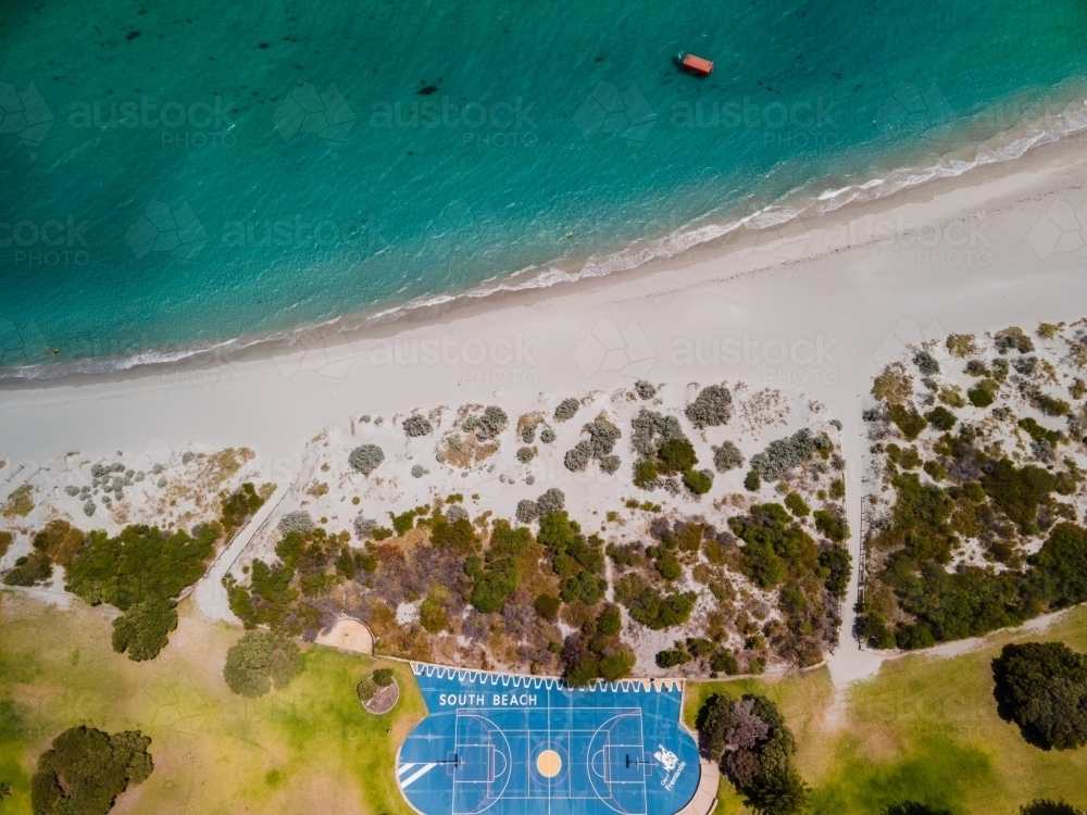 top view shot of a blue basketball court near a beach with white sand, grass plains and green bushes - Australian Stock Image