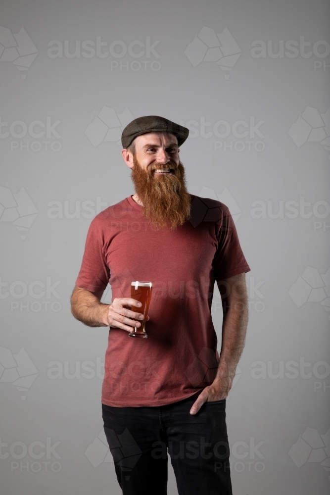 Trendy man with ginger beard and flat cap, holding a glass of beer - Australian Stock Image