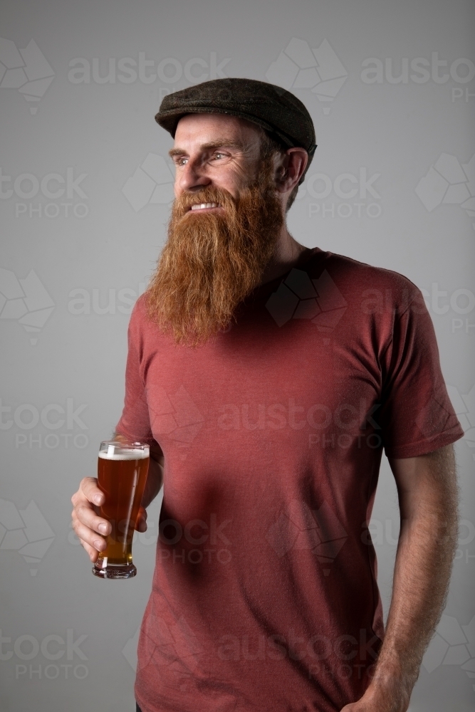 Trendy man with ginger beard and flat cap, holding a glass of beer - Australian Stock Image