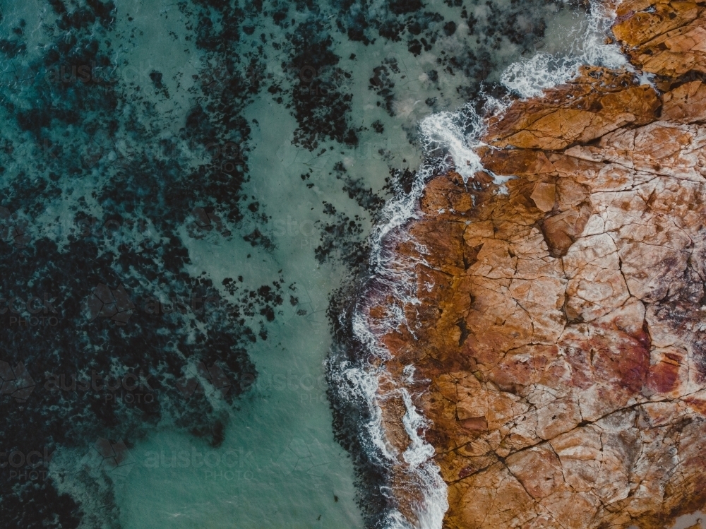 Turquoise blue water crashing against orange rocky shore – abstract aerial - Australian Stock Image