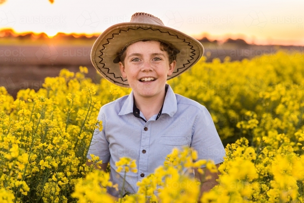 Tween boy smiling wearing hat standing in canola field on farm at sunset - Australian Stock Image