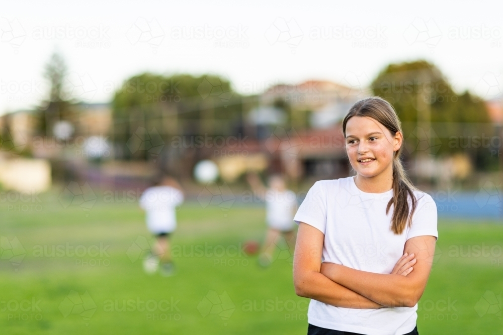 tween girl standing with arms crossed in front of blurry background where other kids play - Australian Stock Image