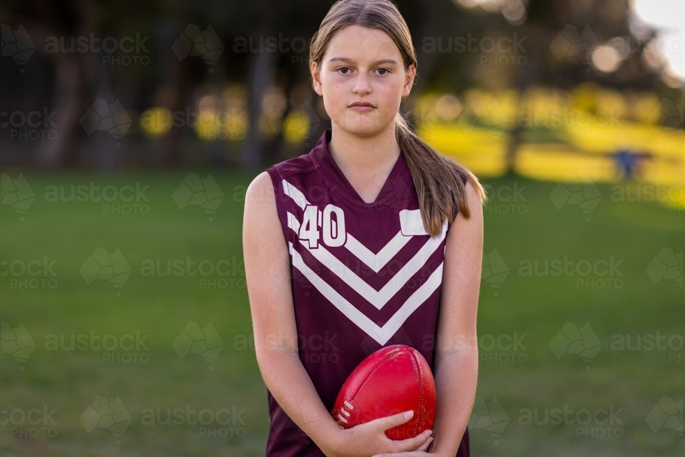 tween girl wearing footy jersey looking at camera holding a red leather football - Australian Stock Image