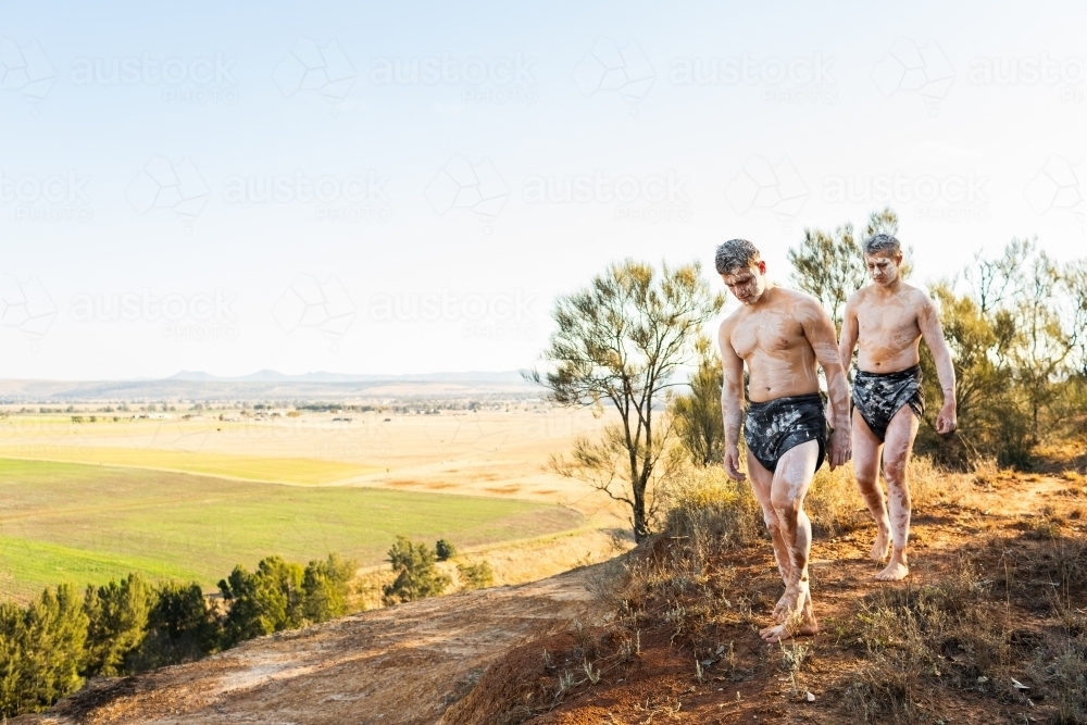 Two Aboriginal Australians on country walking along the edge of a cliff with view over farmland - Australian Stock Image