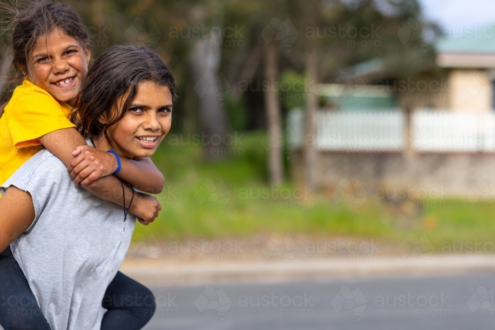 two aboriginal kids piggy-backing outside in rural town - Australian Stock Image