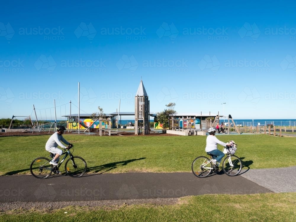 Two cyclists on a cycle path with a children's playground in the background - Australian Stock Image