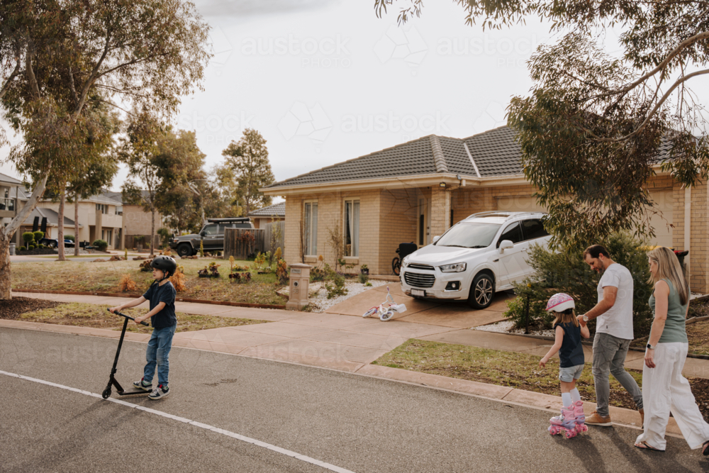 Two kids on the street with their parents one on scooter and the other on roller skates. - Australian Stock Image