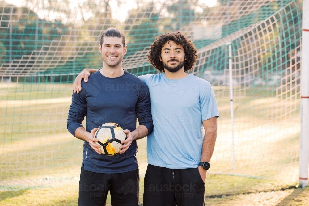 Two men smiling as they stand together in a soccer  goal, with one of the men holding a soccer ball - Australian Stock Image