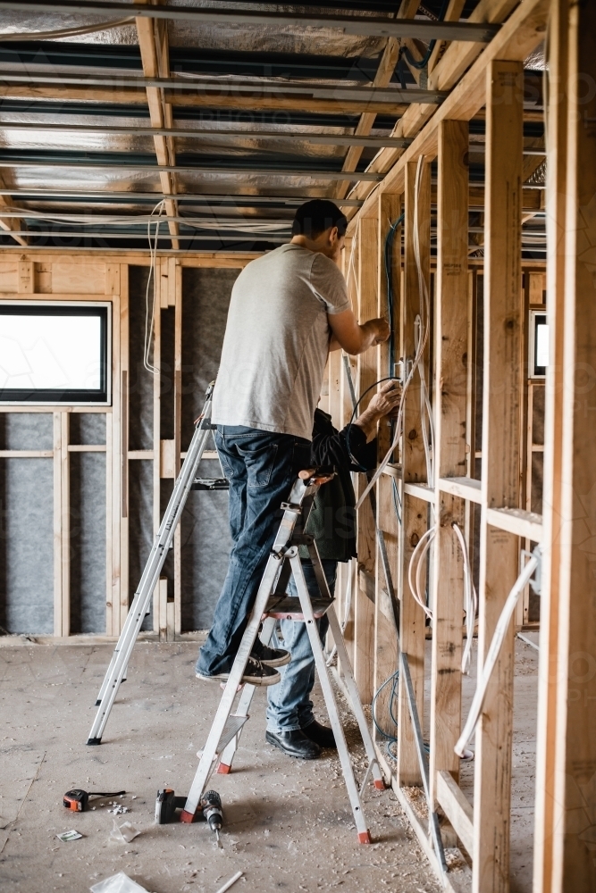 two men, younger on a ladder while older stands, they are laying cables in a house at frame stage - Australian Stock Image