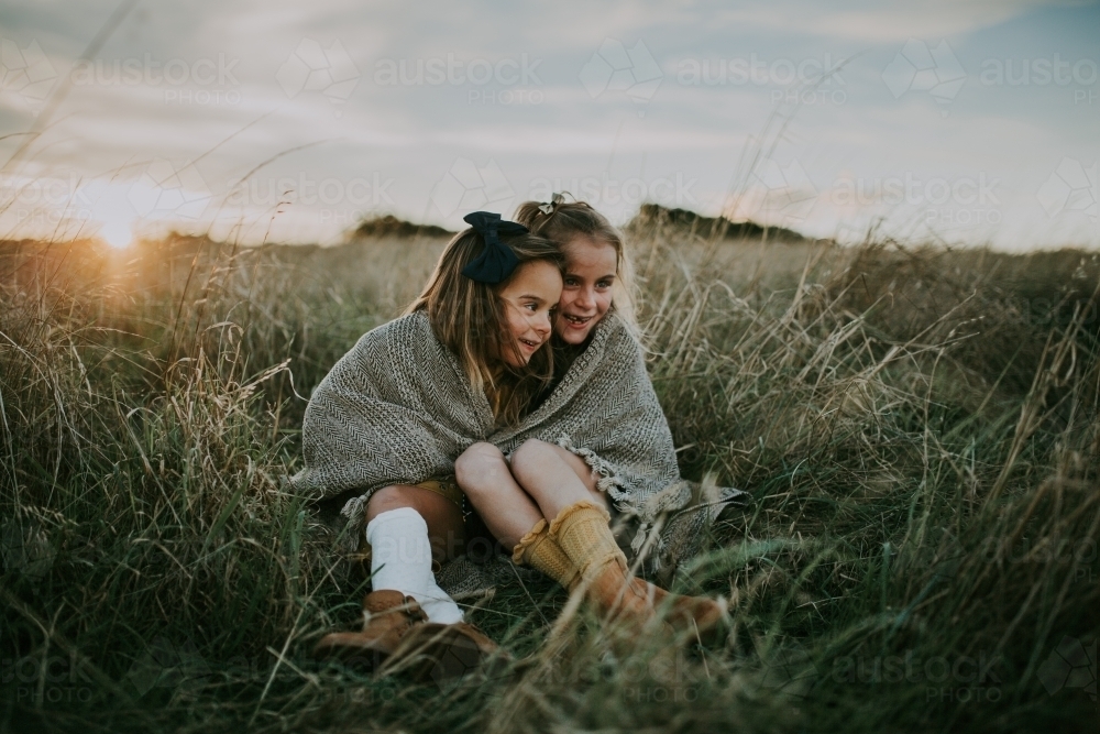 Two sisters sitting in paddock wrapped in blanket at sunset - Australian Stock Image