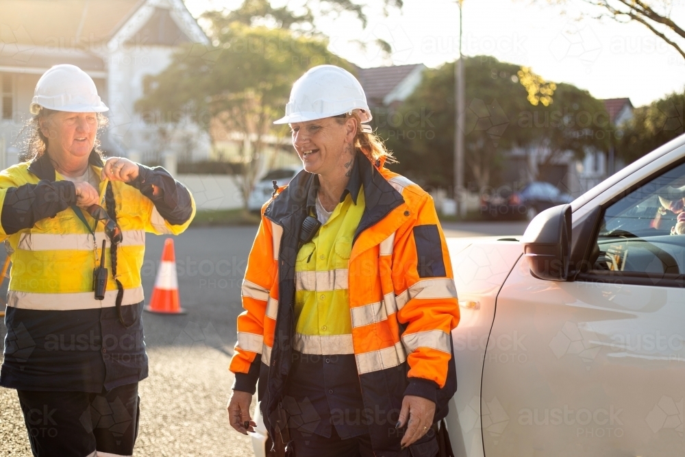 Two smiling women road workers wearing white helmet with orange and yellow jacket on sunset - Australian Stock Image