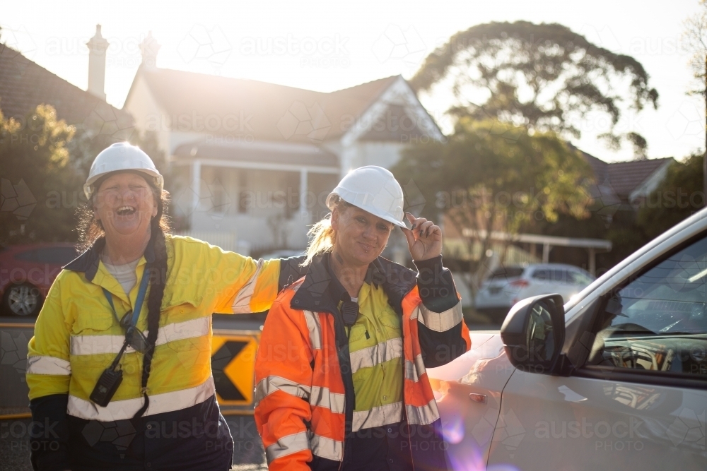 Two smiling women road workers wearing white helmet with orange and yellow jacket on sunset - Australian Stock Image