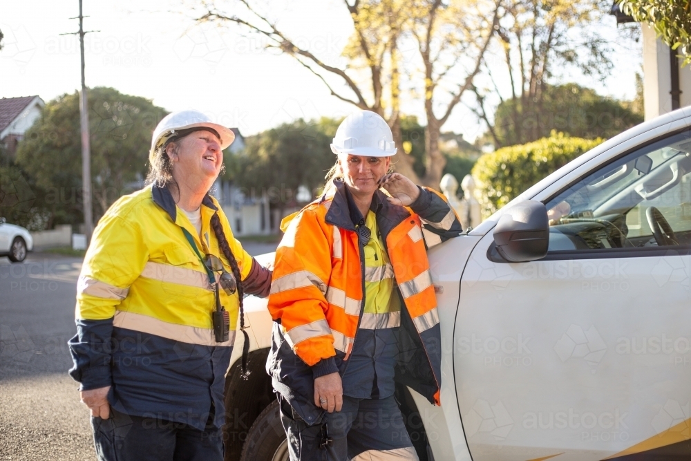 Two smiling women road workers with white helmet leaning on a white car - Australian Stock Image