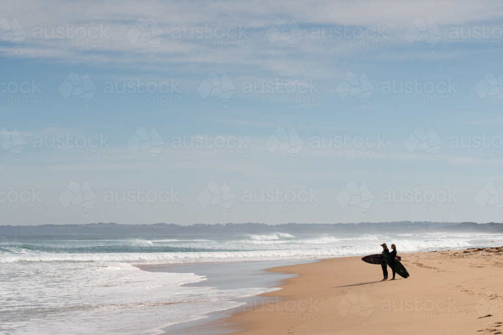 Two surfers with boards standing on beach watching the ocean - Australian Stock Image
