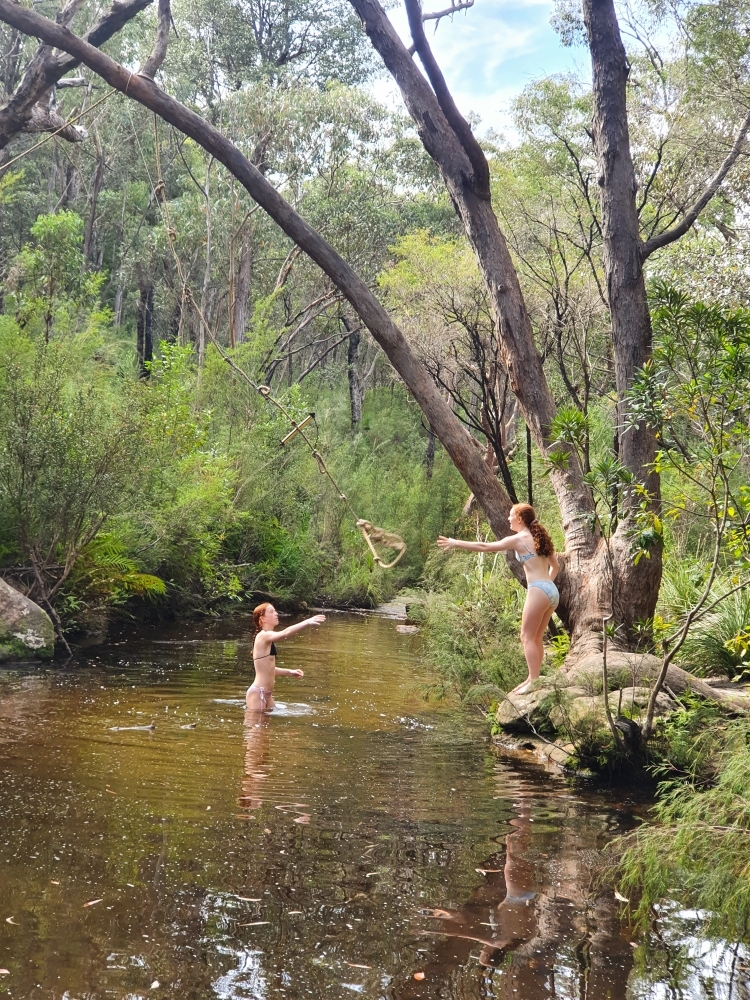 Two teenage girls swinging on a rope swing at a rock pool - Australian Stock Image