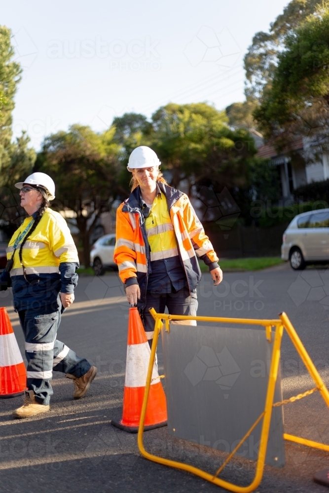 Two women road workers wearing white helmet, yellow and orange jackets on the road with cones - Australian Stock Image