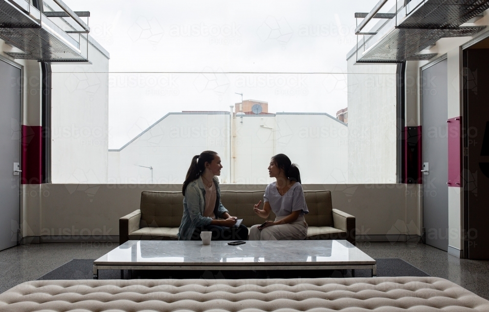Two women sitting and talking to each other on an office sofa near a large window - Australian Stock Image