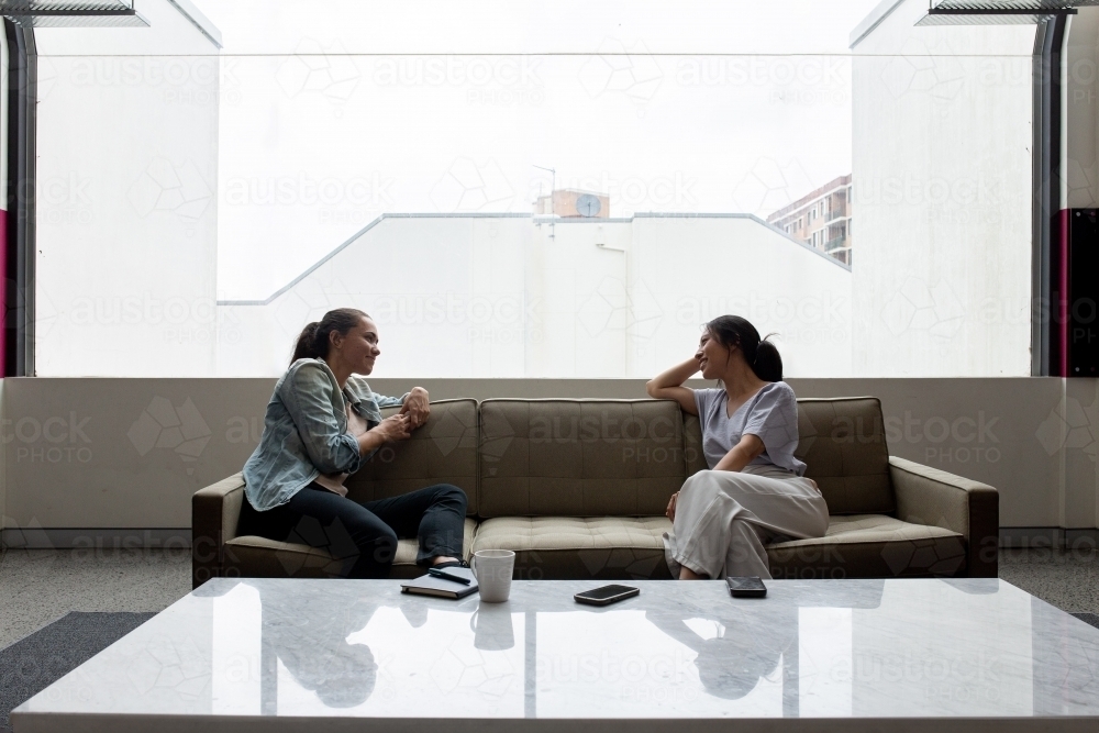 Two women sitting and talking to each other on an office sofa near a large window - Australian Stock Image