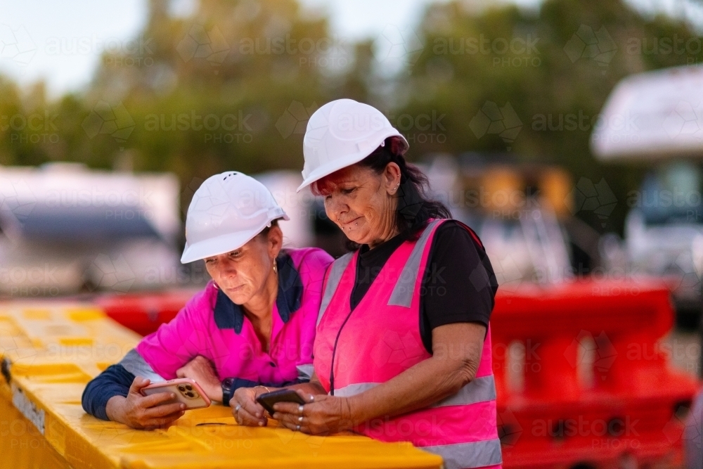 Two women wearing hard hats and pink hi-vis leaning on barrier looking at their phones in low light - Australian Stock Image