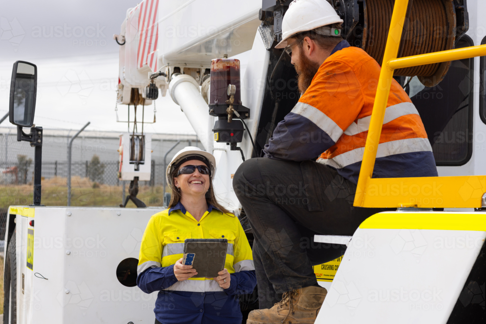 Two workers wearing high visibility clothing man sitting on crane while woman shows him a tablet - Australian Stock Image