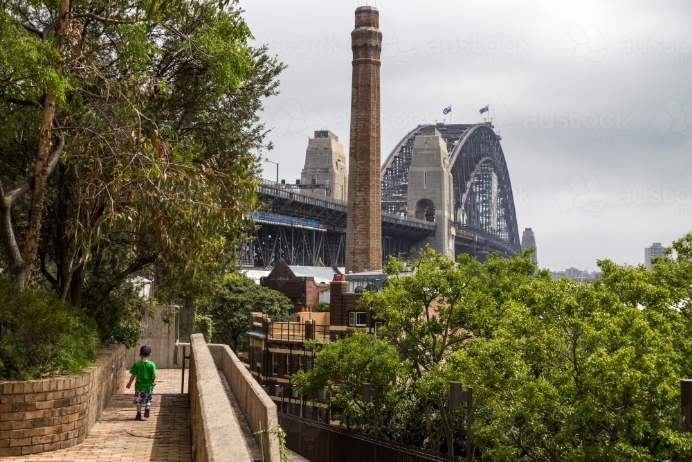 Two year old boy walking along path overlooking foliage with Sydney Harbour Bridge in background - Australian Stock Image