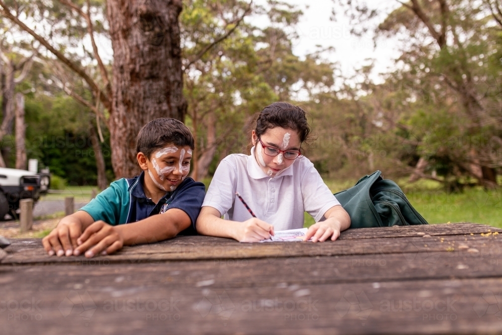 Two young Aboriginal kids wearing white face paints itting at a table drawing - Australian Stock Image