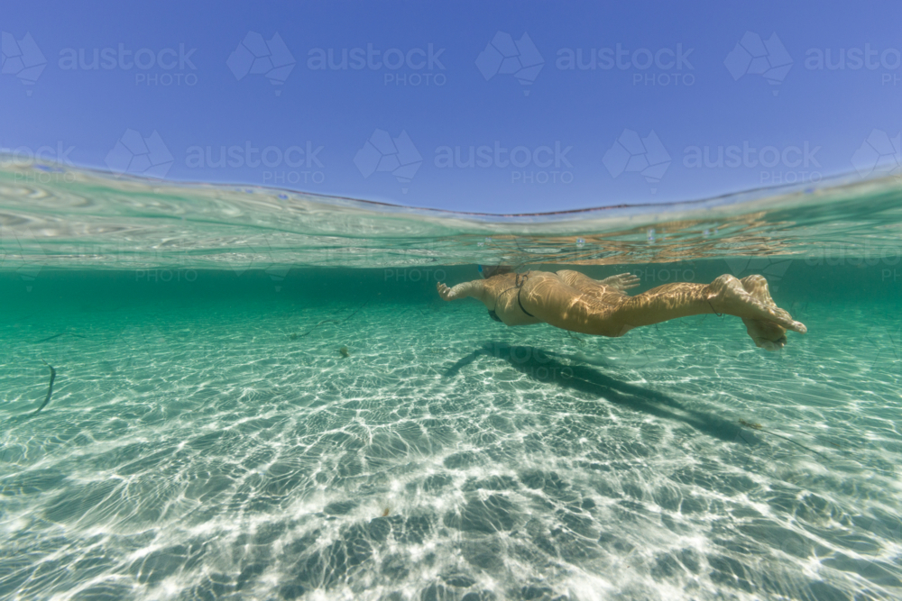Under-over split shot of woman in bikini swimming in clear, turquoise water on summer day - Australian Stock Image