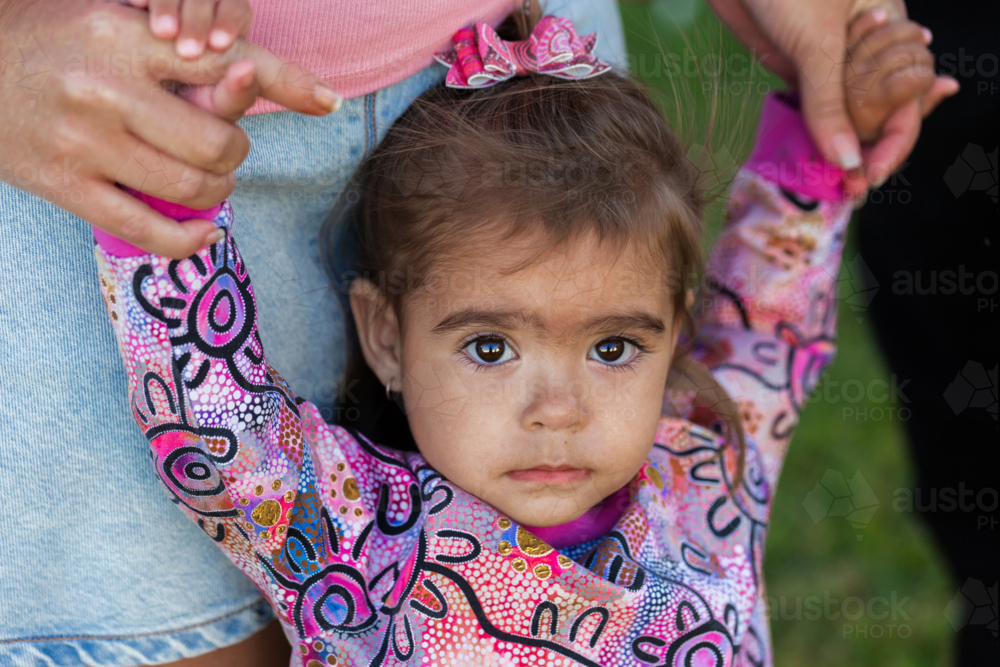 Unsure Aboriginal toddler girl with dark eyes standing close to mum outside - Australian Stock Image