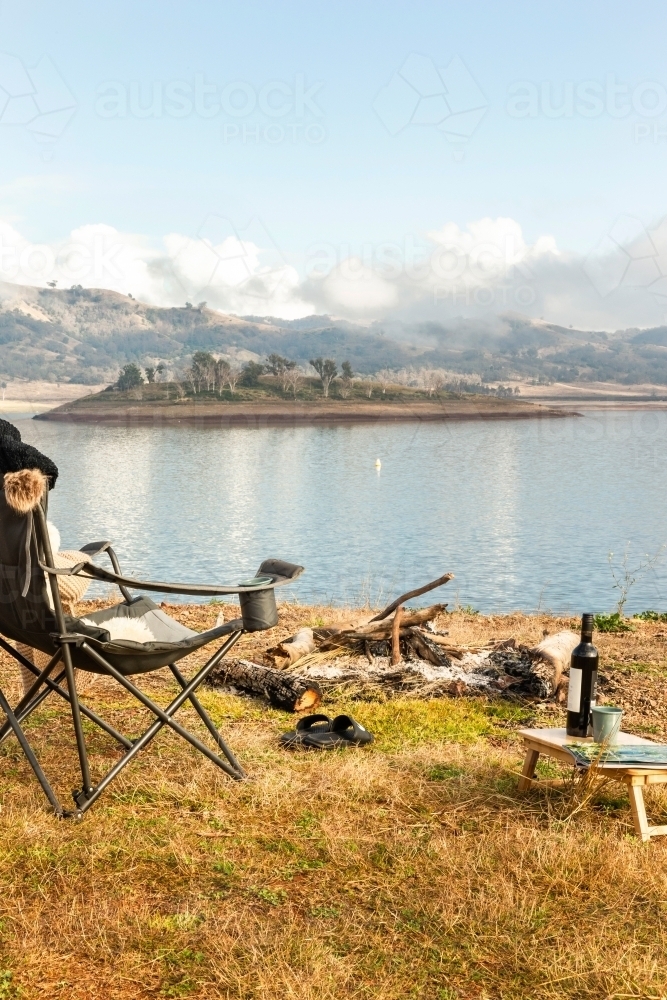 An extinguished campfire with foldable chair pair of slippers and wine beside the lake - Australian Stock Image