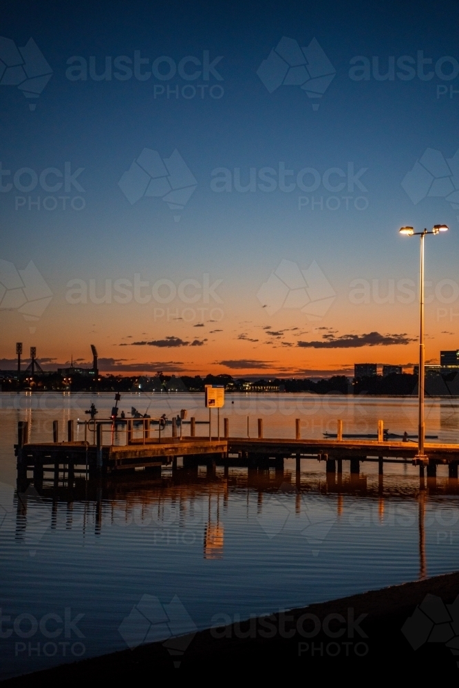 vertical shot of a pier during sunset with lamp post, buildings reflected by the water - Australian Stock Image