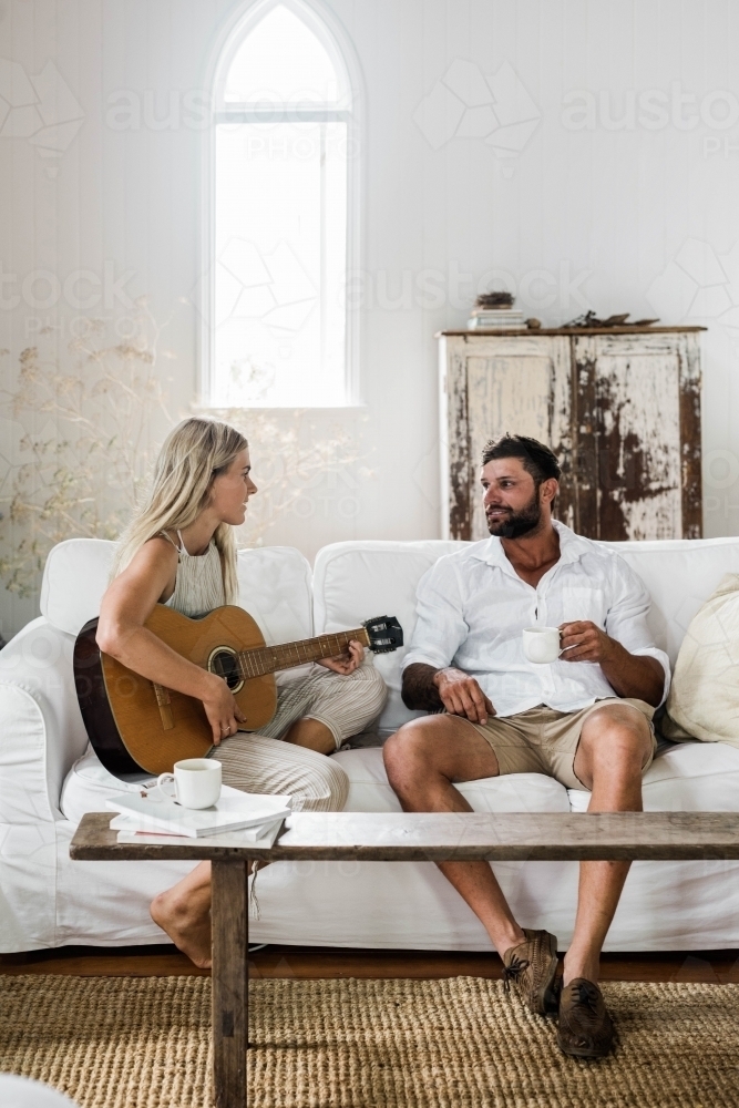 Vertical shot of a woman and a man on a white couch playing guitar and drinking coffee - Australian Stock Image