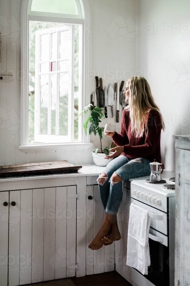 Vertical shot of a woman sitting on the kitchen counter near the window while drinking coffee - Australian Stock Image