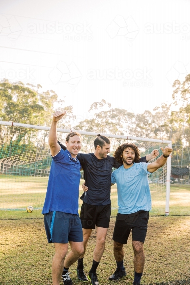 vertical shot of men smiling while raising their arms in the field with goal in the background - Australian Stock Image