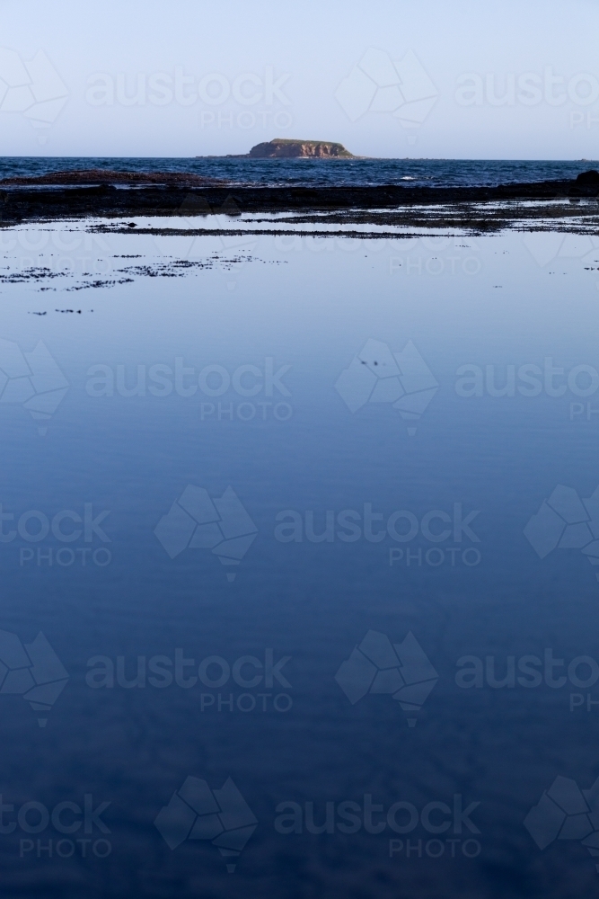 vertical shot of the ocean with sky reflecting on it and a mountain island at the back - Australian Stock Image