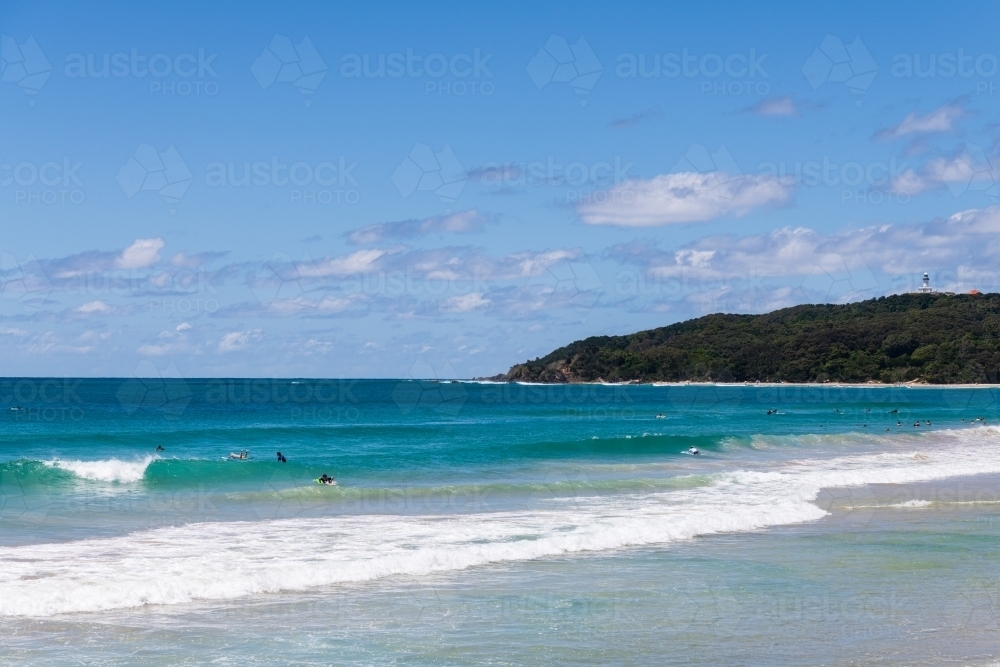 View across the water to Cape Byron and lighthouse from  Byron Bay with surfers - Australian Stock Image