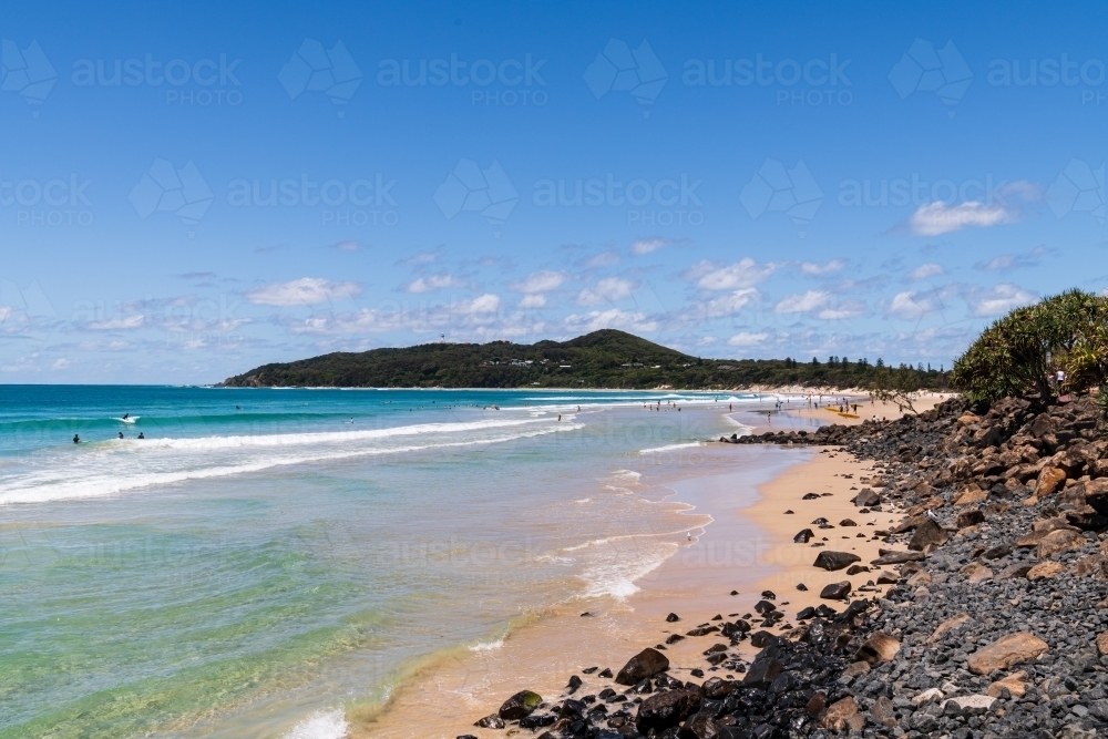 View of Cape Byron and lighthouse from the beach at Byron Bay with surf, rocks and sandy foreshore - Australian Stock Image