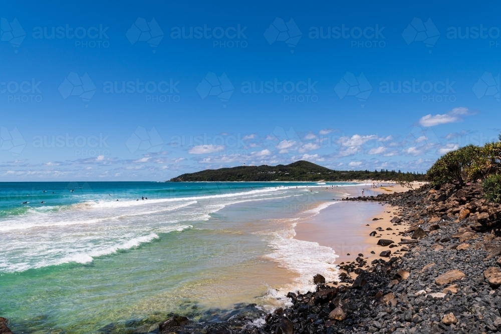 View of Cape Byron and lighthouse from the beach at Byron Bay with surfers rocks and sandy foreshore - Australian Stock Image