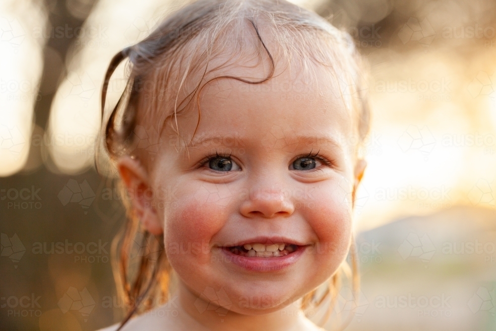 Wet grinning little kid in afternoon sunset light after playing in sprinkler water - Australian Stock Image