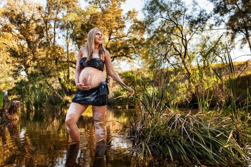 Wide shot of Aboriginal Australian woman standing in creek water in traditional body paint - Australian Stock Image