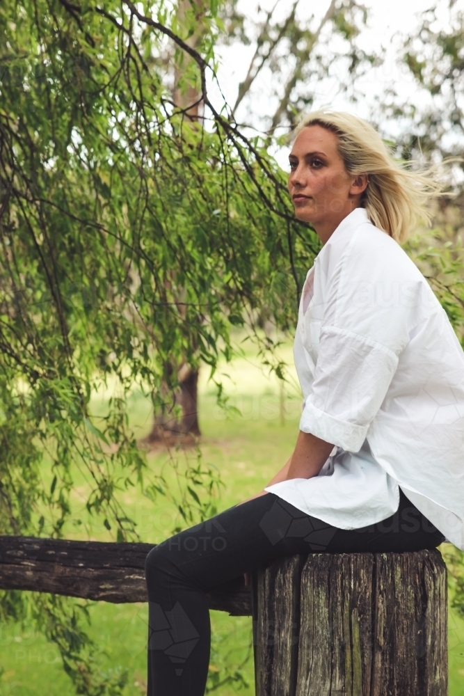 Wind blowing young woman's blonde hair as she sits on a fence post - Australian Stock Image