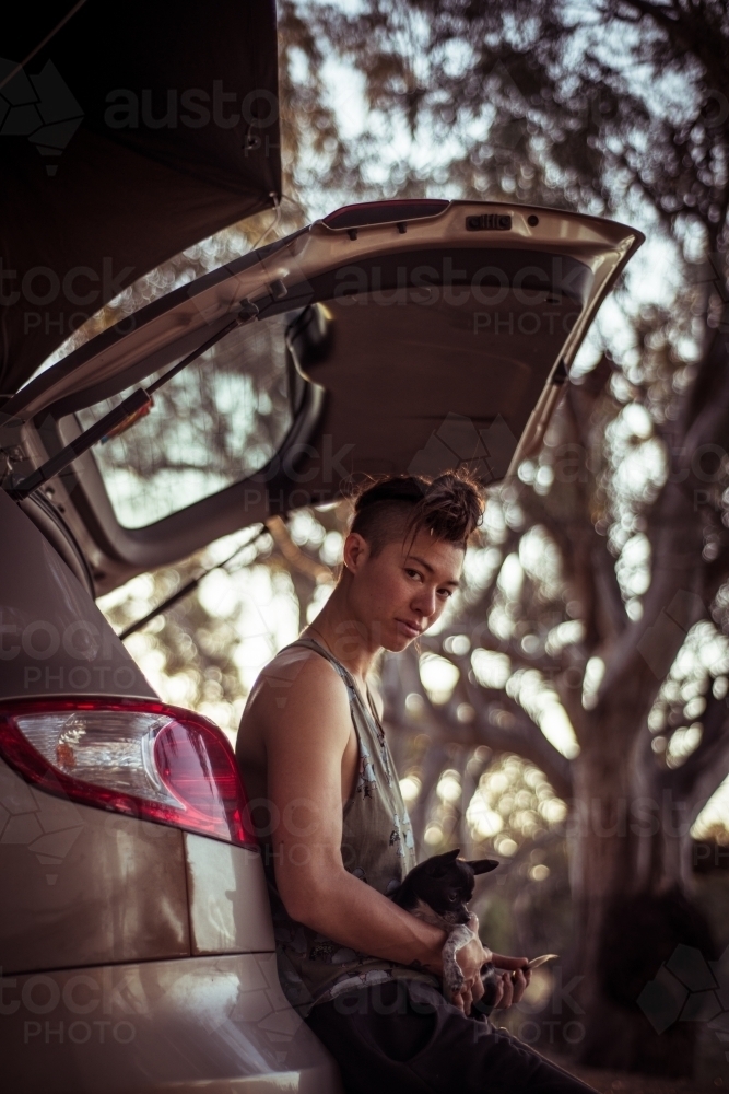 Woman holding a dog as she leans on the back of a car in the bush - Australian Stock Image