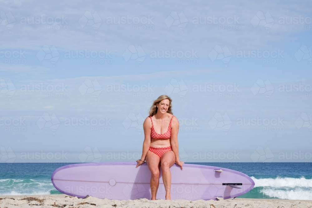 Woman in bikini sitting on longboard surfboard on shoreline of beach - Australian Stock Image