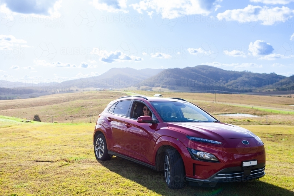 Woman in electric car in country paddock location with hills - Australian Stock Image