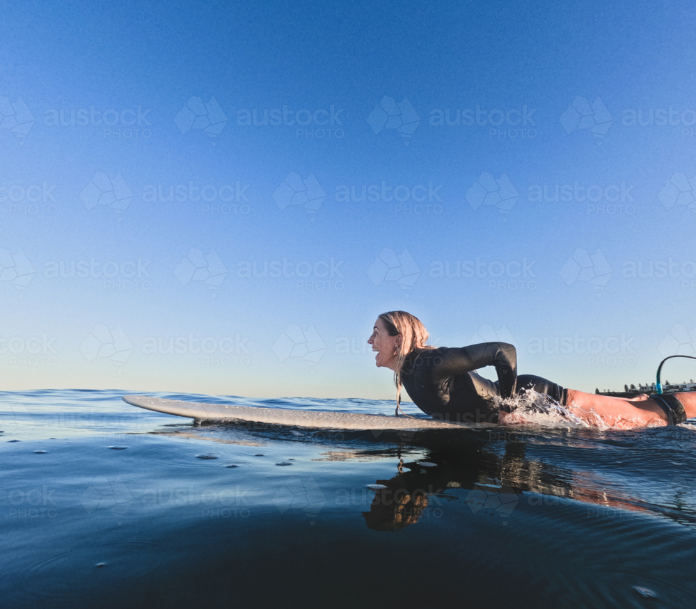 Woman in summer wetsuit lying on longboard surfboard paddling looking out to glassy sea laughing - Australian Stock Image
