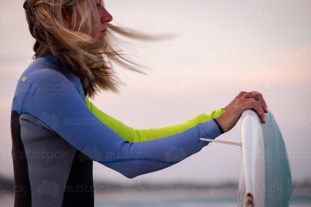 Woman in wetsuit holding surfboard on beach with windswept hair - Australian Stock Image