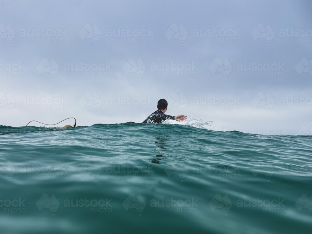 Woman looking away paddling surfboard across ocean taken at water level with overcast sky - Australian Stock Image