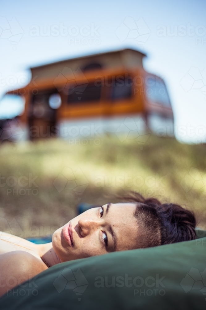 Woman lying with head on pillows outdoors with camper van in background - Australian Stock Image