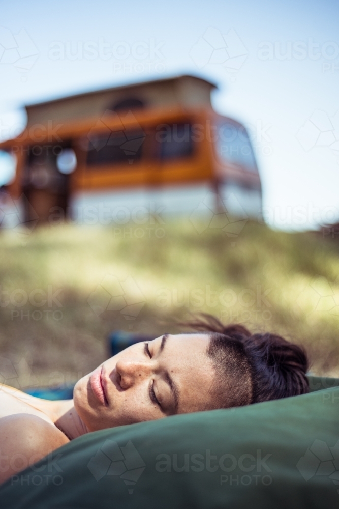 Woman lying with head on pillows outdoors with camper van in background - Australian Stock Image