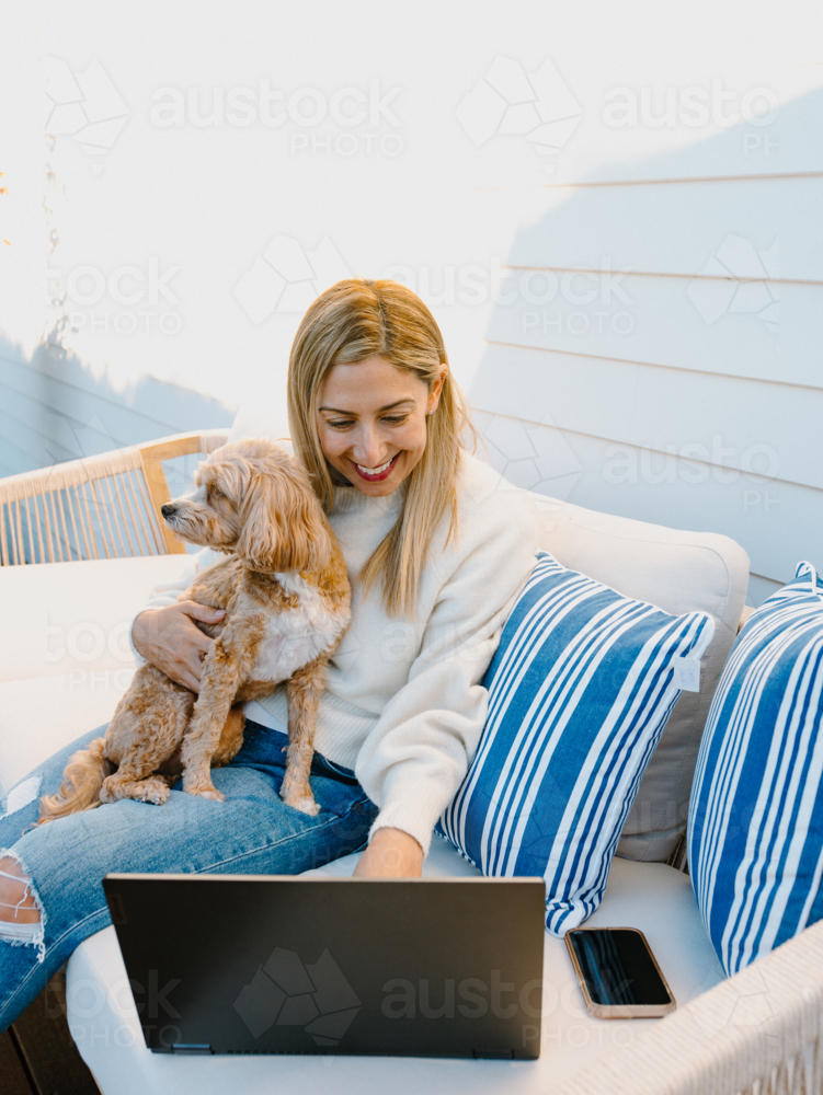 Woman on her laptop while holding a dog on the couch. - Australian Stock Image