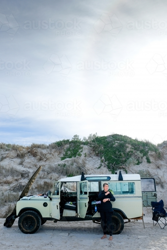 Woman putting on wetsuit on beach in front of vintage Land Rover with surfboard leaning on car - Australian Stock Image